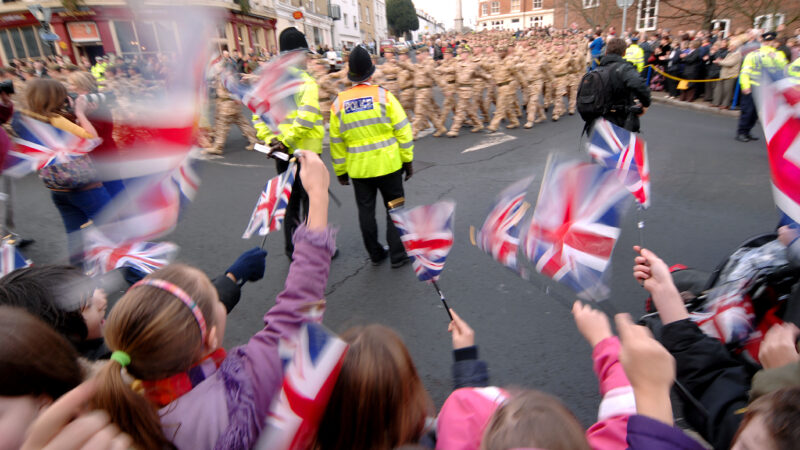 Children waving flags during a military parade, with police standing on the side of the road.